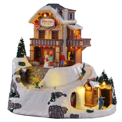 Villaggio natalizio Lemax Winterhaus Resort illuminato, con chalet innevato, sciatori, snowborder e impianto di risalita.