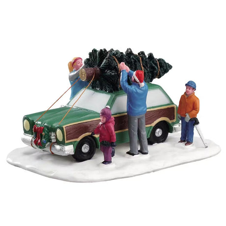 Famiglia che carica un albero di Natale sul tetto di un'auto verde parcheggiata su base innevata.