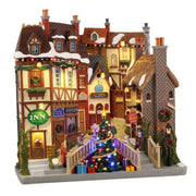 Facciata natalizia in stile inglese con locanda, pub, libreria e al centro un albero di Natale illuminato con pacchi regalo.