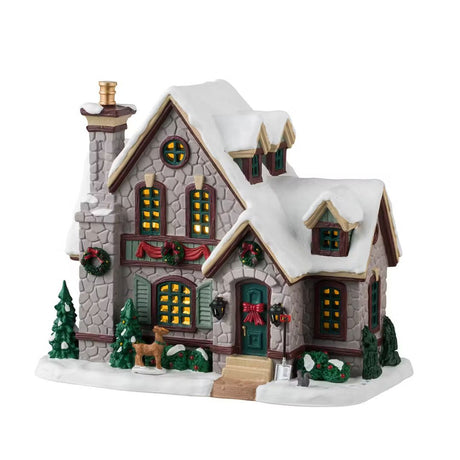 Casetta natalizia illuminata con tetto innevato, ghirlande verdi e fiocco rosso sulla porta, decorata con alberelli e renna in miniatura.
