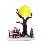 Babbo Natale e Signora Claus seduti su una panchina sotto un albero spoglio illuminato da una luna gialla.