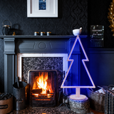 Albero di Natale in legno sbiancato con illuminazione LED blu, posizionato accanto a un camino acceso.