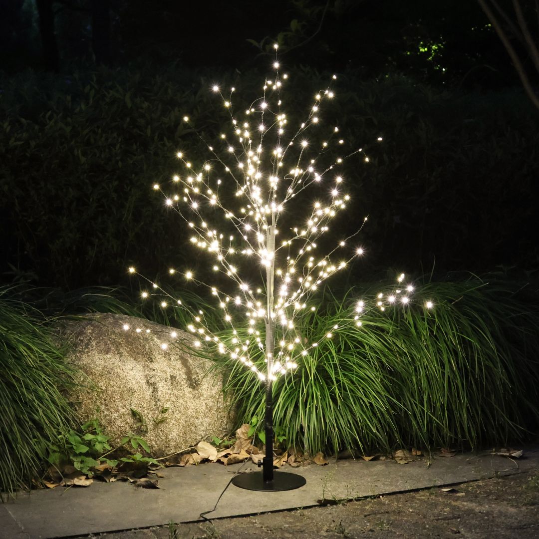 Albero decorativo a LED bianco caldo da esterno acceso di notte, posizionato su una base circolare.