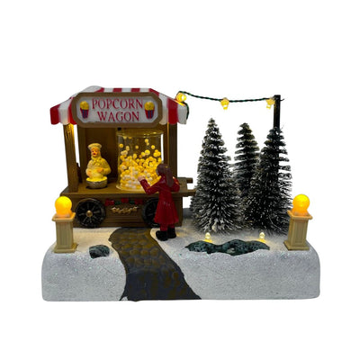 Miniatura natalizia con bancarella Popcorn Wagon, personaggio in cappotto rosso e alberi innevati decorativi.