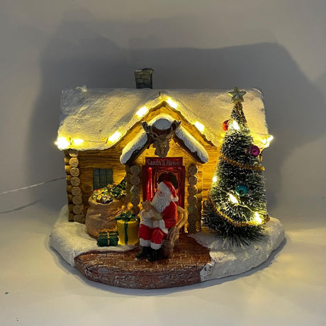 Casetta natalizia innevata con Babbo Natale seduto all’ingresso, albero decorato e pacchi regalo davanti alla porta con luci LED.