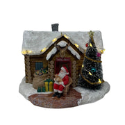 Casetta natalizia innevata con Babbo Natale seduto all’ingresso, albero decorato con luci LED e pacchi regalo davanti alla porta.