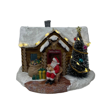 Casetta natalizia innevata con Babbo Natale seduto all’ingresso, albero decorato con luci LED e pacchi regalo davanti alla porta.