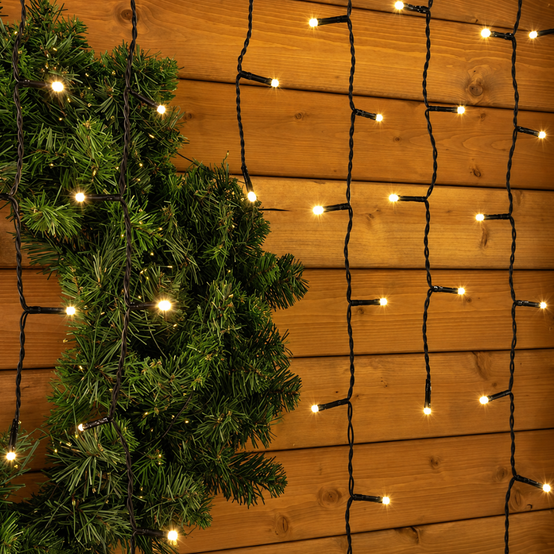 Dettaglio della tenda luminosa Ice Light con LED bianco caldo, cavo nero e rami di abete su parete in legno.
