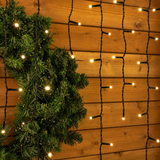 Dettaglio della tenda luminosa Ice Light con LED bianco caldo, cavo nero e rami di abete su parete in legno.
