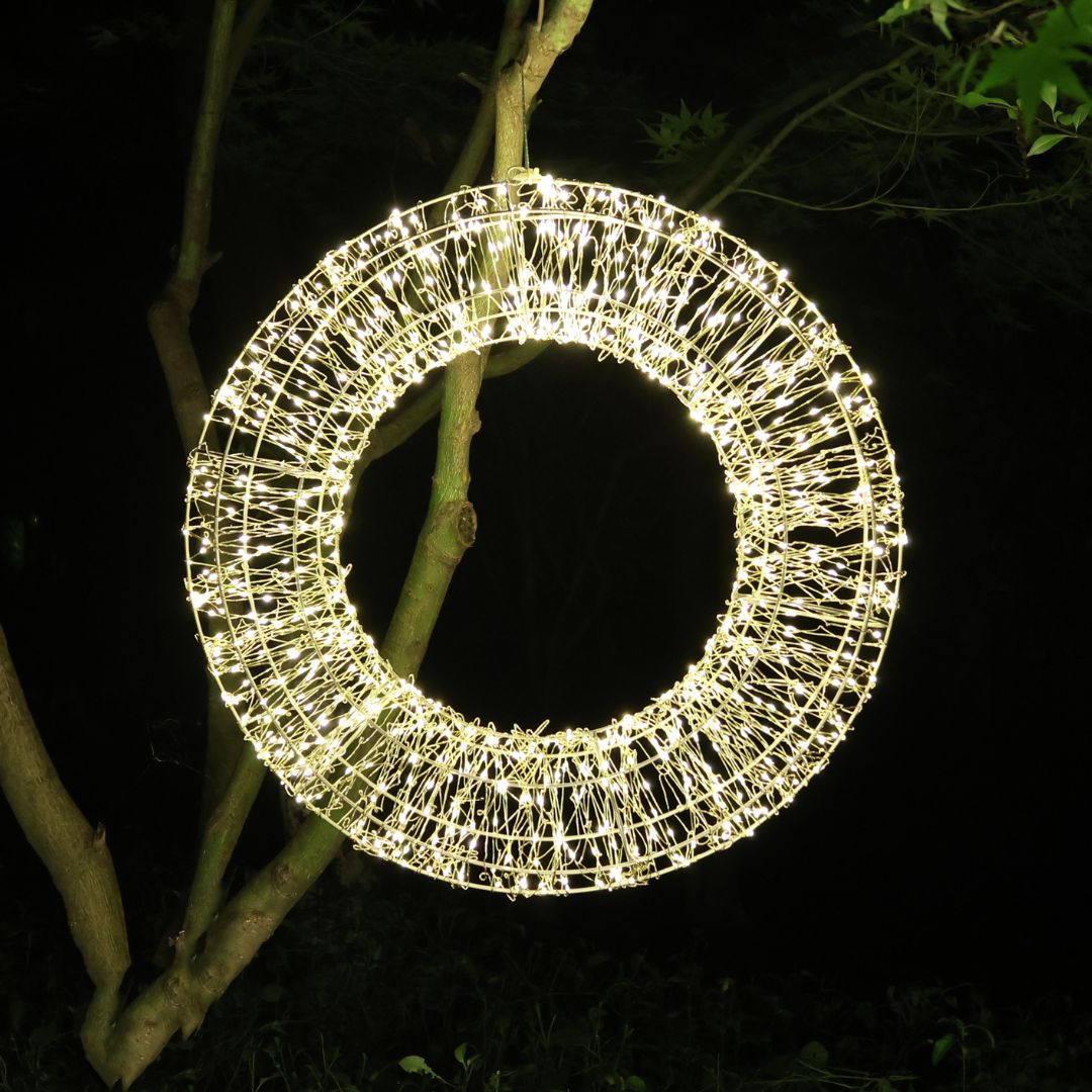 Corona luminosa decorativa a LED bianco caldo appesa a un albero in esterno durante la notte.