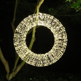 Corona luminosa decorativa a LED bianco caldo appesa a un albero in esterno durante la notte.