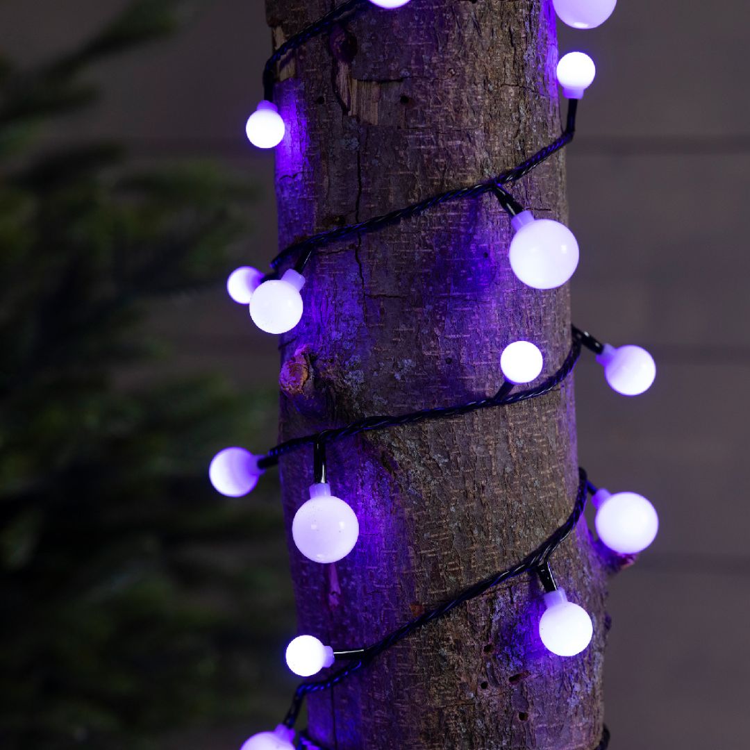 Luci LED multicolore a forma di ciliegia, avvolte attorno al tronco di un albero, illuminate di color lilla.
