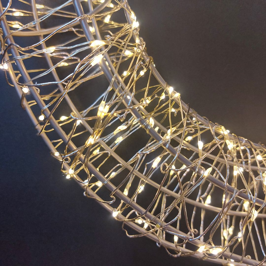 Dettaglio della corona luminosa decorativa con fili metallici intrecciati e micro LED a luce bianco caldo.