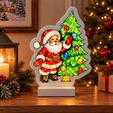 Decorazione luminosa con Babbo Natale che addobba l’albero, ambientata su un mobile tra luci e decorazioni natalizie