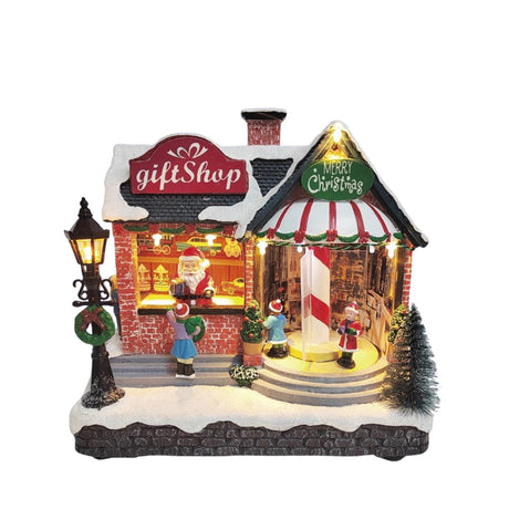 Negozio natalizio in miniatura con insegna gift shop, vetrina illuminata con Babbo Natale e bambini che osservano decorazioni sotto la neve su sfondo bianco.