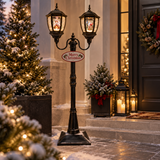 Lampione natalizio doppio nero illuminato, ambientato all'ingresso di una casa decorata per Natale con alberi innevati e lanterne