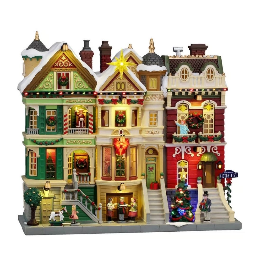 Facciata Lemax con eleganti case vittoriane innevate e illuminate, decorate con ghirlande, festoni e luci natalizie, con personaggi e albero di Natale lungo la strada.