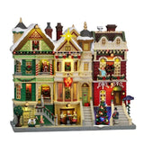 Facciata Lemax con eleganti case vittoriane innevate e illuminate, decorate con ghirlande, festoni e luci natalizie, con personaggi e albero di Natale lungo la strada.
