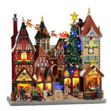 Grande scena natalizia Lemax con edifici in stile tradizionale, bancarelle illuminate, albero di Natale decorato, torre con orologio e slitta di Babbo Natale con renne sullo sfondo.