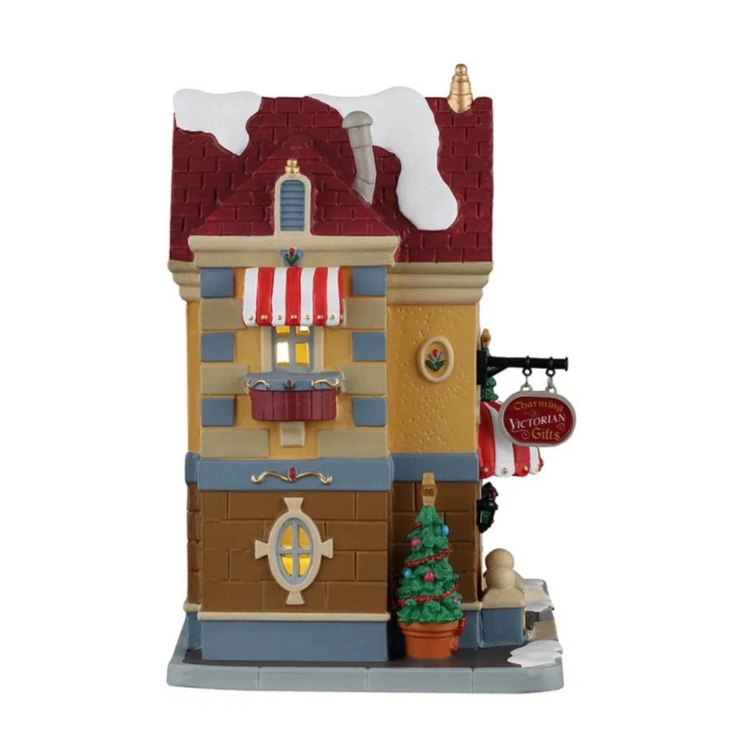 Father Christmas Gift Shop Lemax visto di lato, con finestra illuminata, tenda a righe rosse e bianche, insegna “Charming Victorian Gifts” e piccolo albero di Natale decorato.