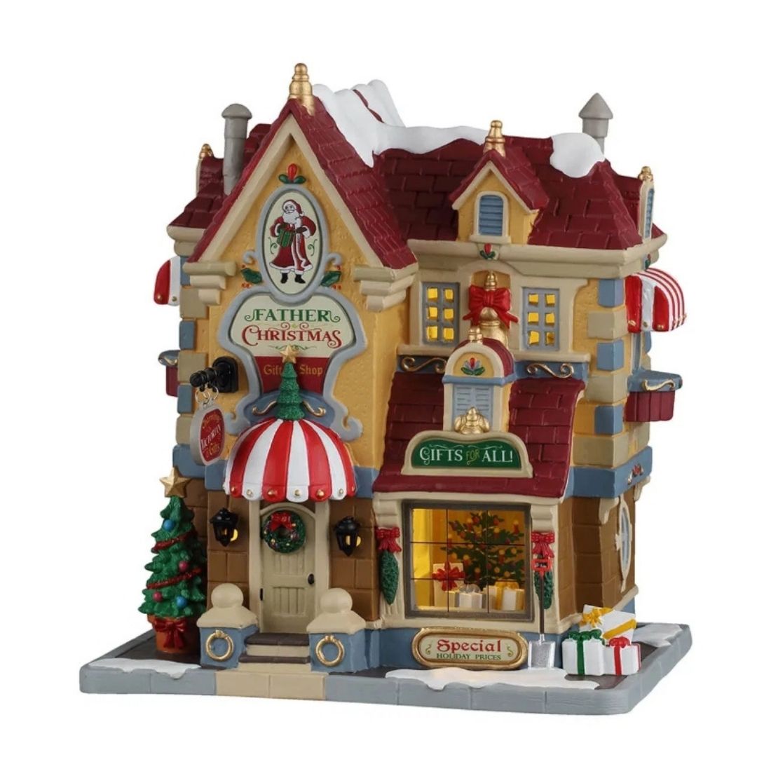 Father Christmas Gift Shop Lemax, negozio di regali natalizi innevato e illuminato, con insegne decorative, vetrina con pacchi regalo, albero di Natale e dettagli rossi e oro.