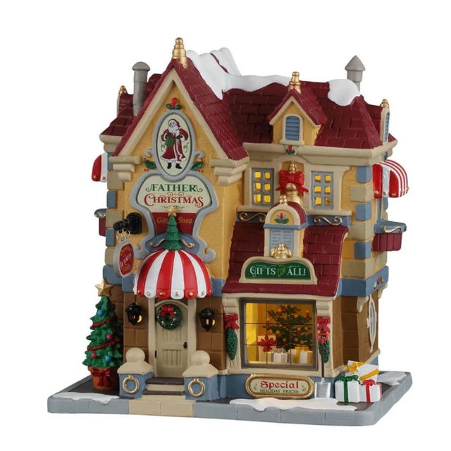 Father Christmas Gift Shop Lemax, negozio di regali natalizi innevato e illuminato, con insegne decorative, vetrina con pacchi regalo, albero di Natale e dettagli rossi e oro.