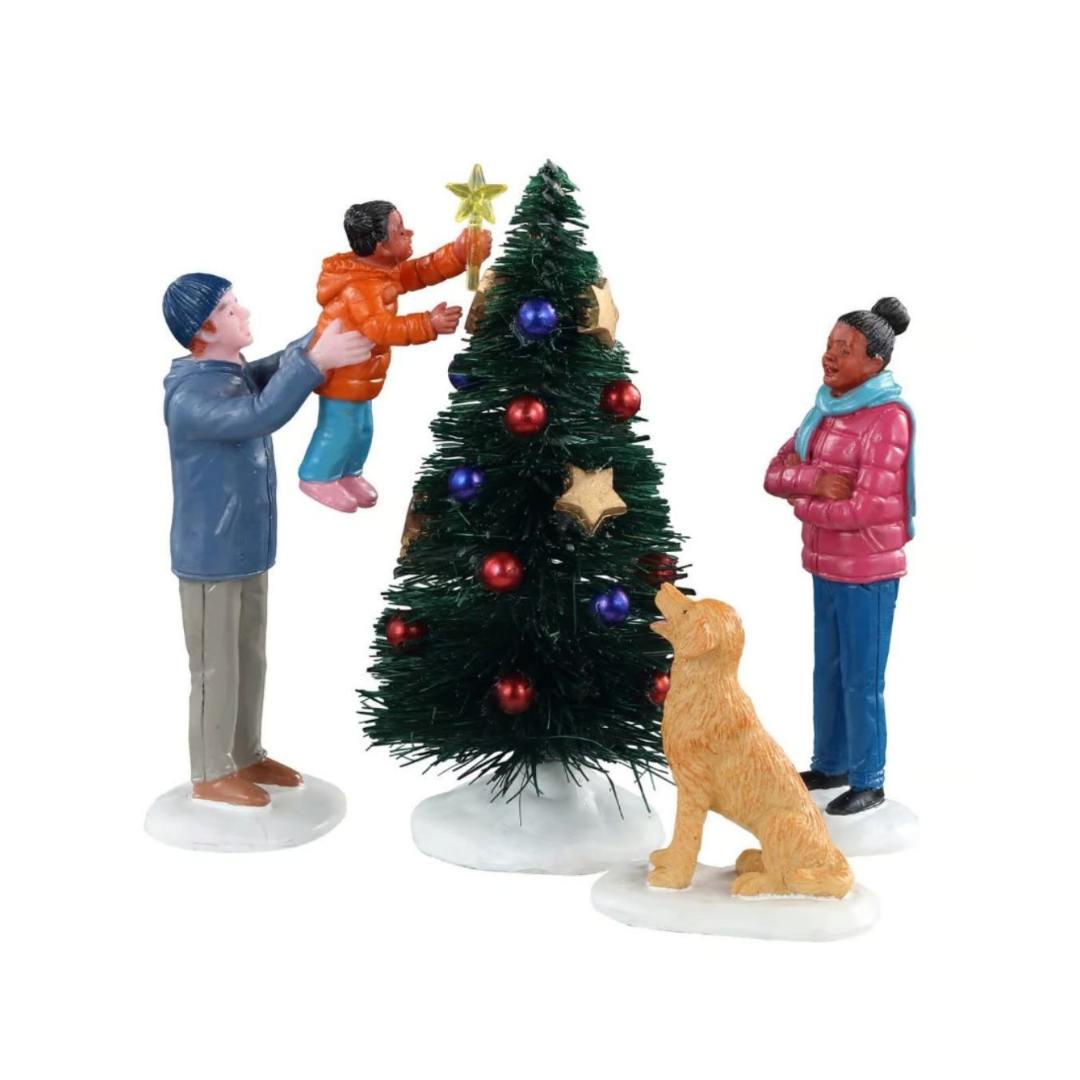 Final Touch Lemax con famiglia che decora l’albero di Natale: adulto solleva un bambino per posizionare la stella in cima, mentre una donna e un cane osservano la scena.