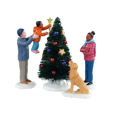 Final Touch Lemax con famiglia che decora l’albero di Natale: adulto solleva un bambino per posizionare la stella in cima, mentre una donna e un cane osservano la scena.