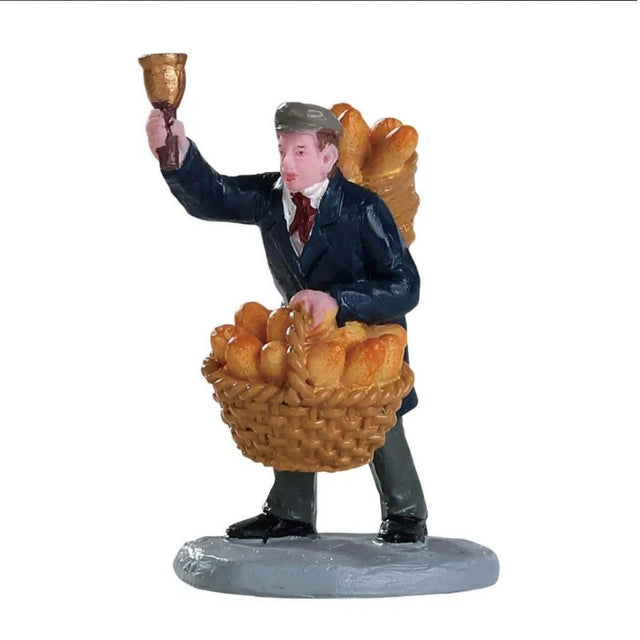 Venditore di pane Lemax con cesta piena di pagnotte e baguette, mentre alza una campanella per richiamare i clienti.