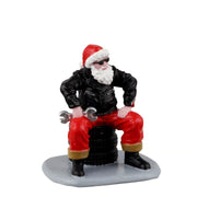 Babbo Natale in giacca nera, occhiali da sole e pantaloni rossi, seduto su una pila di pneumatici con una chiave inglese.
