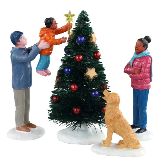 Final Touch Lemax con famiglia che decora l’albero di Natale: adulto solleva un bambino per posizionare la stella in cima, mentre una donna e un cane osservano la scena.
