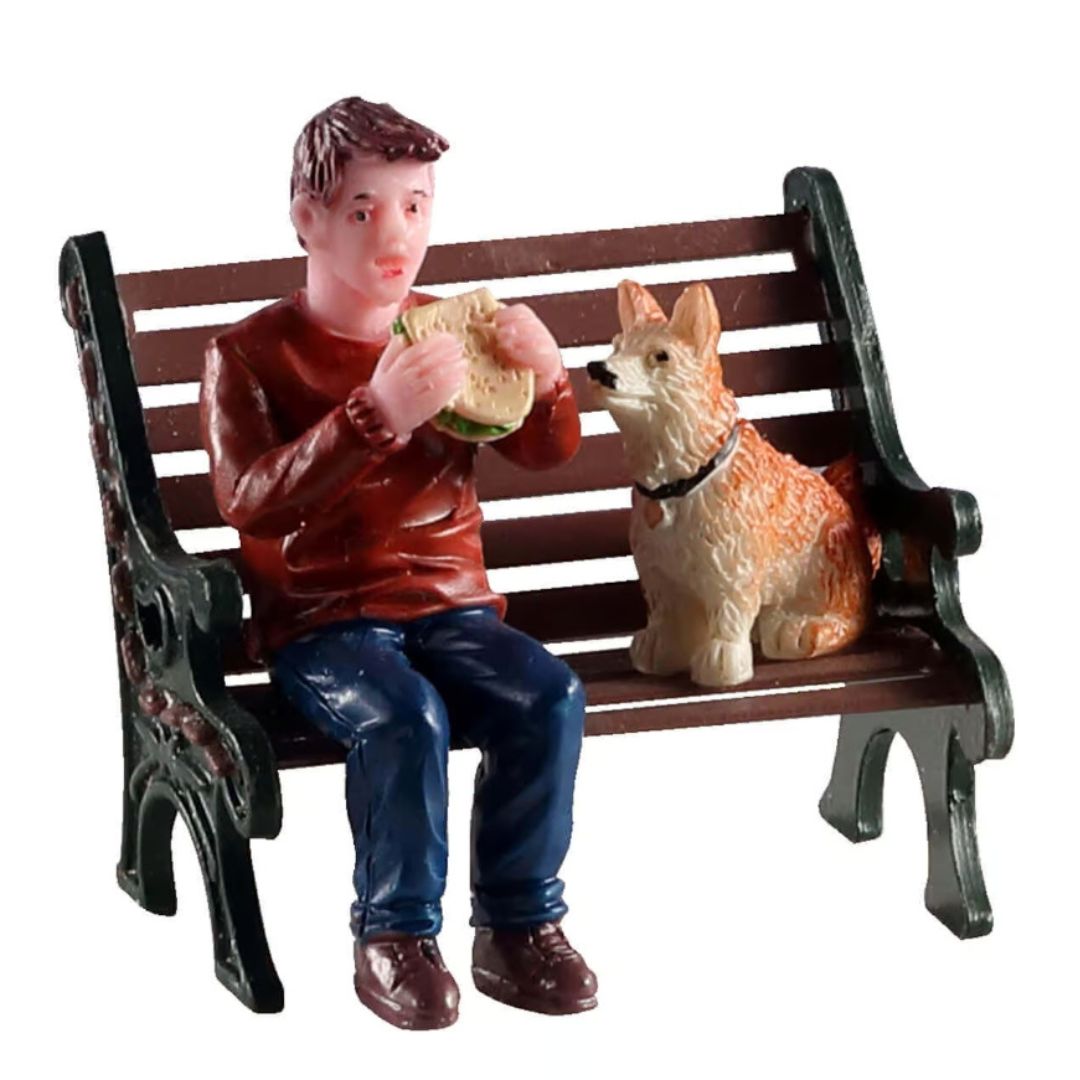 Personaggi Lemax Lunchbreak Buddy con ragazzo seduto su una panchina mentre mangia un panino, accanto al suo cane che lo osserva.