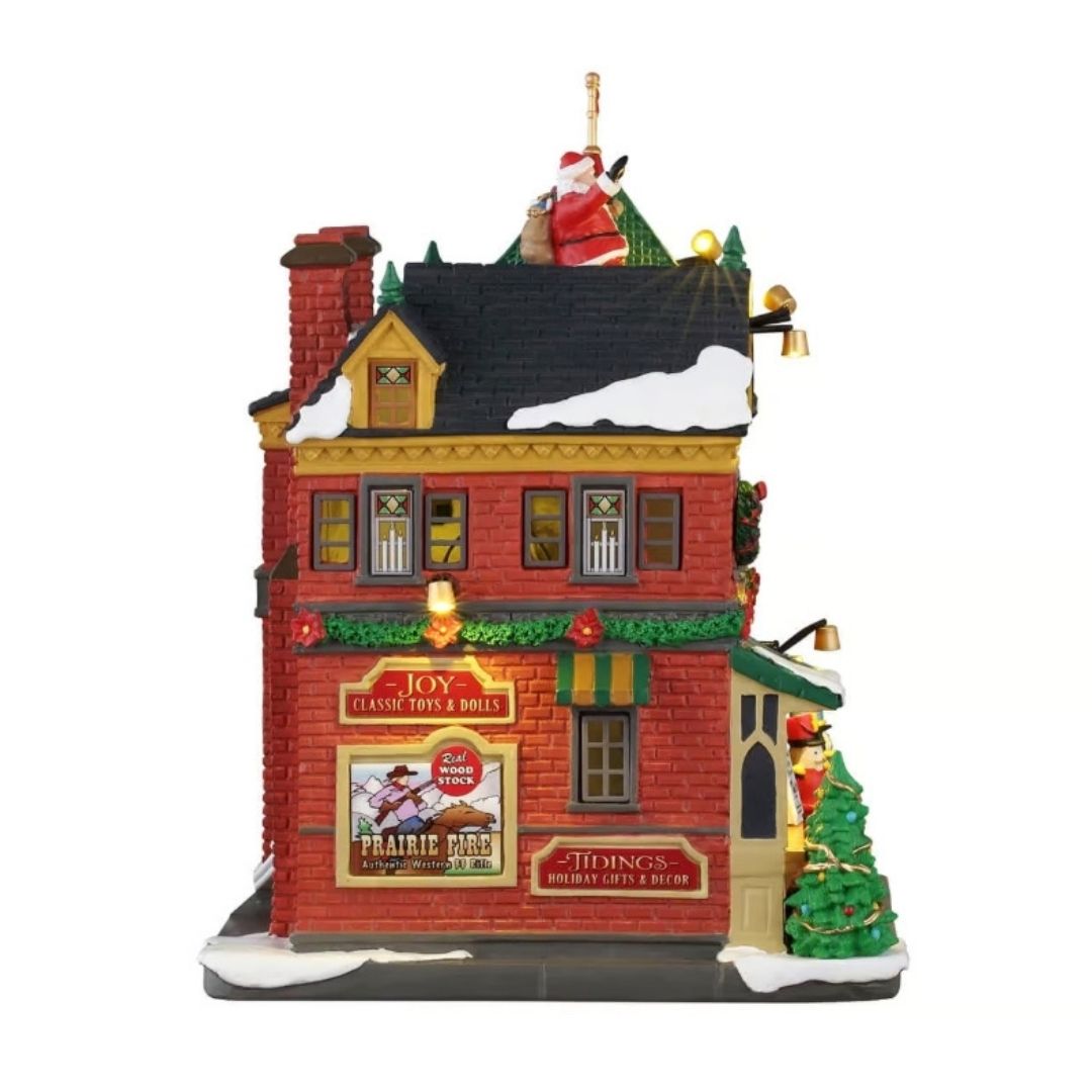 Tidings and Joy Toy Shop Lemax visto di lato, con pareti in mattoni rossi, ghirlande natalizie, insegne, tetto innevato e Babbo Natale sul tetto.