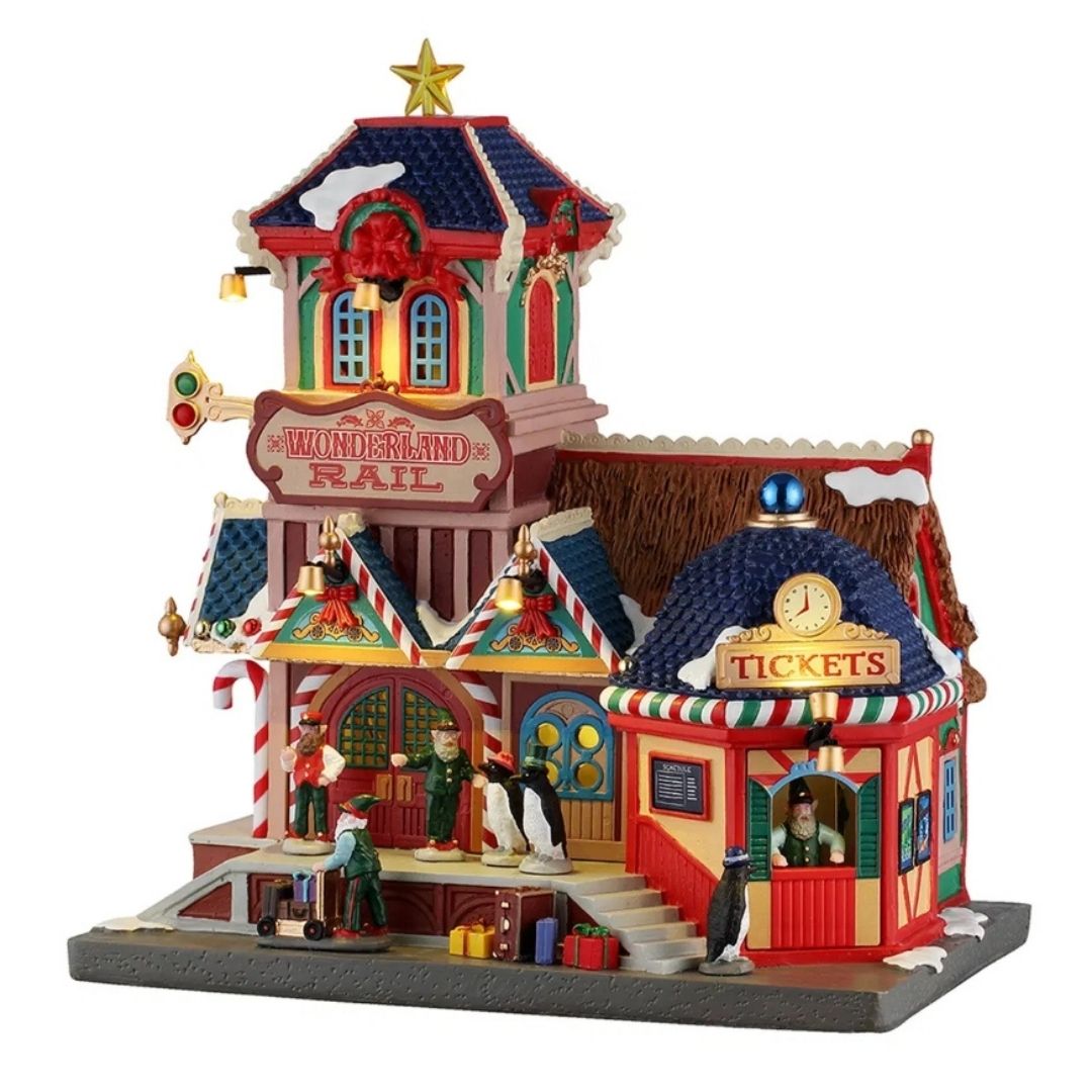 Stazione ferroviaria natalizia Lemax innevata e illuminata, con biglietteria, torre con stella, personaggi, pinguini, valigie e dettagli decorativi.