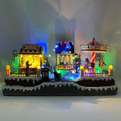 Villaggio natalizio luminoso a tema luna park con giostra, bancarella di popcorn e chiosco del caffè, decorato con luci LED multicolore e personaggi in miniatura.