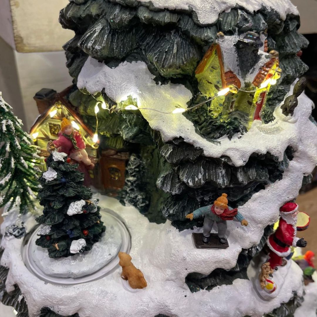 Scena ravvicinata del villaggio natalizio con albero decorato, figure di Babbo Natale e bambini che giocano sulla neve.