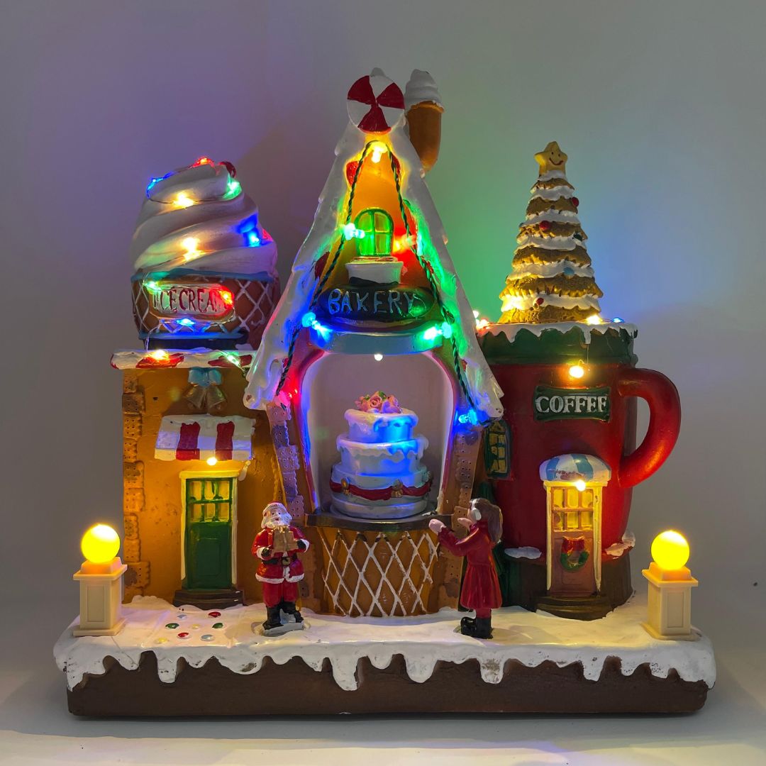 Negozio di dolci natalizio luminoso con casette a tema pasticceria, gelateria e caffè, decorato con luci LED multicolore e figure di Babbo Natale e una bambina.
