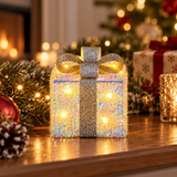Pacco regalo luminoso da 13 cm con fiocco argentato, ambientato su un mobile tra decorazioni natalizie illuminate