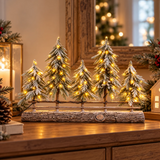 Cinque pini di Natale innevati con luci LED bianco caldo e staccionata, ambientati su un mobile in legno decorato per Natale.