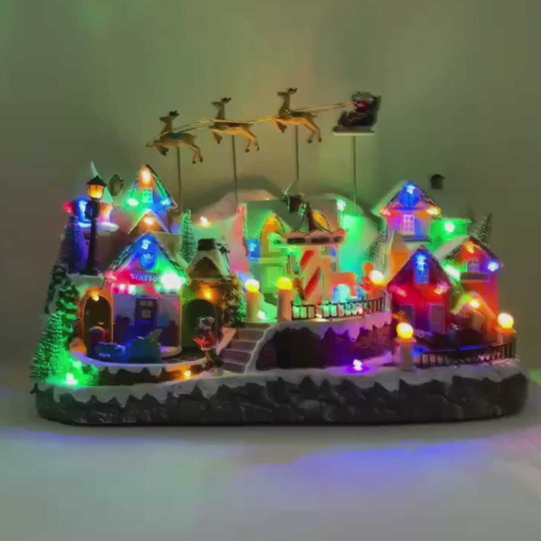Video del villaggio natalizio con case innevate, giostra, trenino e slitta di Babbo Natale trainata da renne, illuminato da luci LED multicolore.