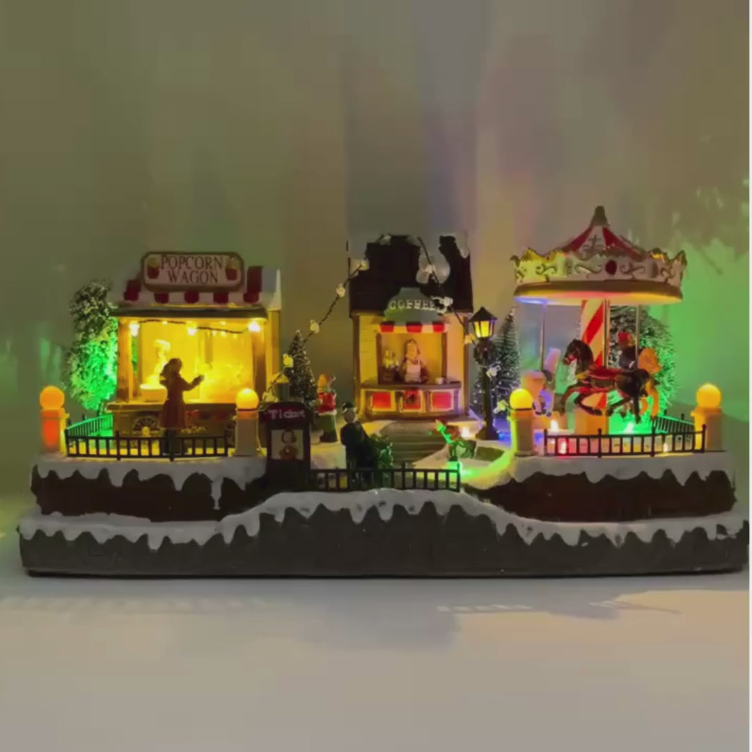 Video del villaggio natalizio a tema luna park con giostra, bancarella di popcorn e chiosco del caffè, decorato con luci LED multicolore.