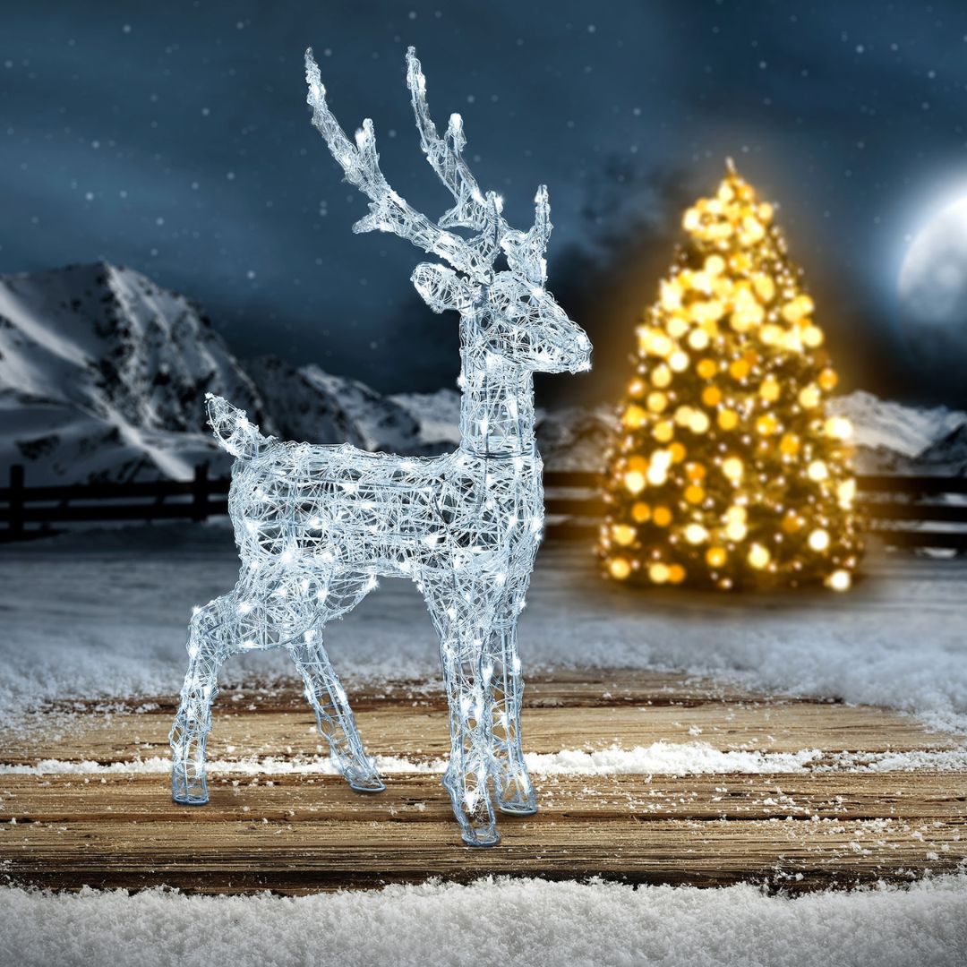 Renna luminosa 85 cm in acrilico posizionata su un pavimento in legno all’aperto, con sfondo innevato, montagne e albero di Natale illuminato sullo sfondo.
