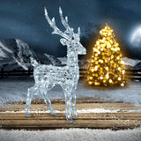 Renna luminosa 85 cm in acrilico posizionata su un pavimento in legno all’aperto, con sfondo innevato, montagne e albero di Natale illuminato sullo sfondo.