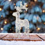 Renna luminosa in acrilico trasparente su tavola innevata con albero decorato sullo sfondo.