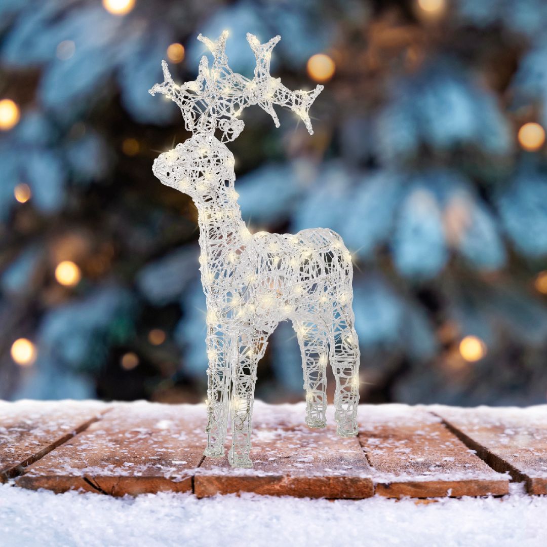 Figura natalizia a forma di renna in acrilico con LED, posizionata su tavola innevata con sfondo natalizio.