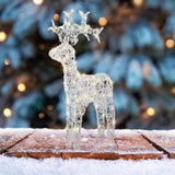 Figura natalizia a forma di renna in acrilico con LED, posizionata su tavola innevata con sfondo natalizio.