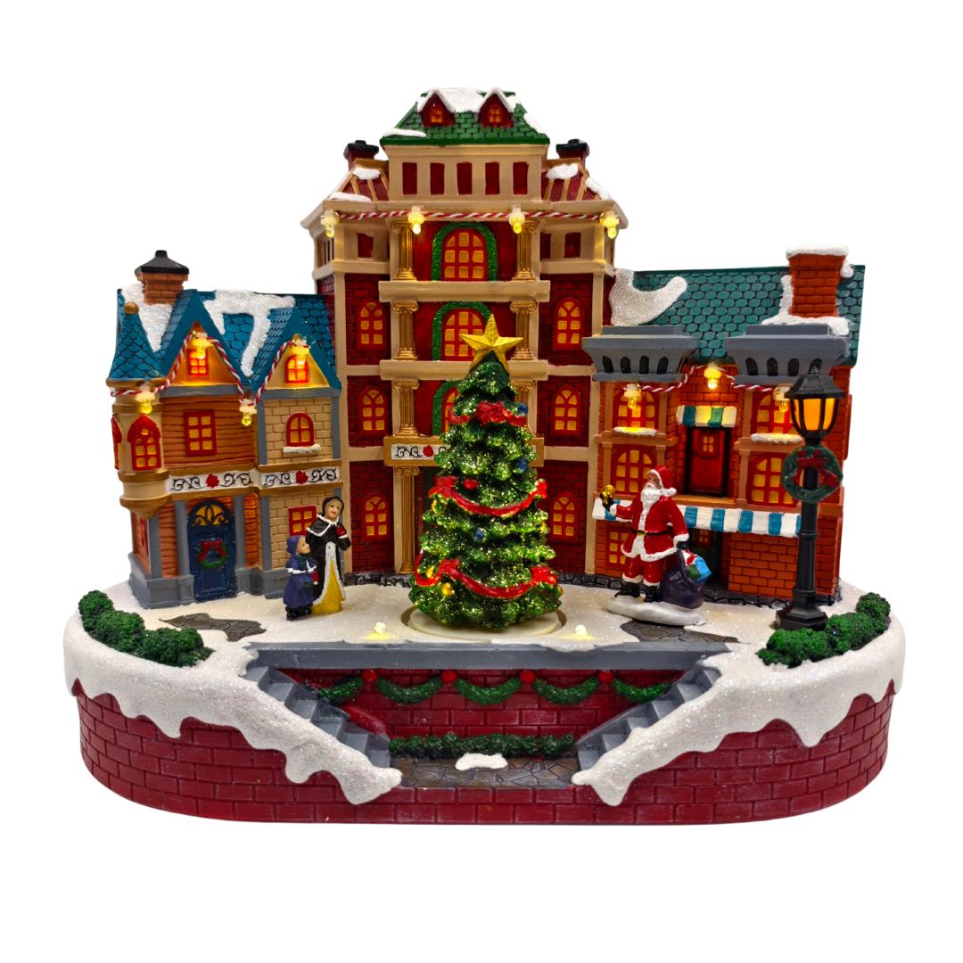 Scena natalizia in miniatura con palazzi illuminati, grande albero decorato al centro, figure di persone e Babbo Natale in una piazza innevata.