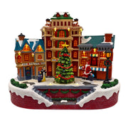 Scena natalizia in miniatura con palazzi illuminati, grande albero decorato al centro, figure di persone e Babbo Natale in una piazza innevata.