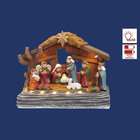 Scena della Natività con statuine colorate, capanna in legno e luci LED calde alla base su sfondo blu.