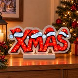 Scritta luminosa XMAS con cappelli di Babbo Natale, ambientata su un mobile accanto ad albero e decorazioni natalizie